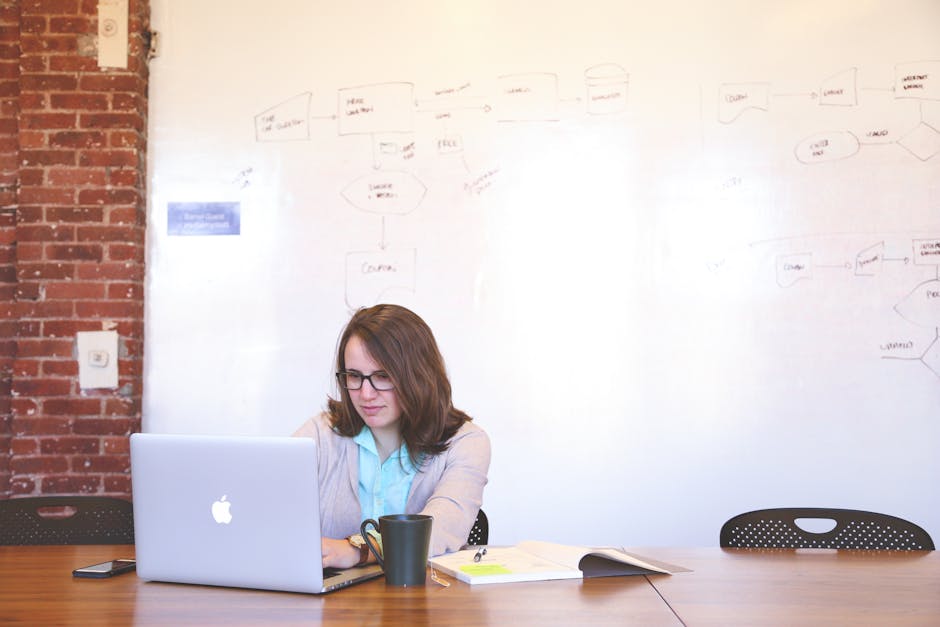 Professional woman using a laptop at her workspace with a whiteboard background.